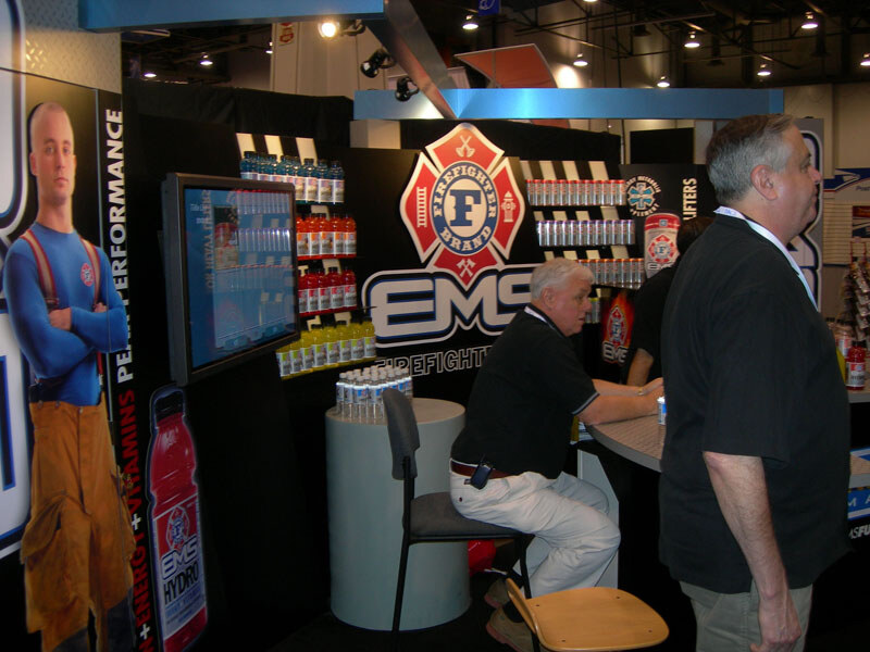 At a black EMS Firefighter Brand booth with a fire department-style logo and shelves of bottles, one man sits at a counter while another man stands in the foreground; a firefighter figure is on the left.