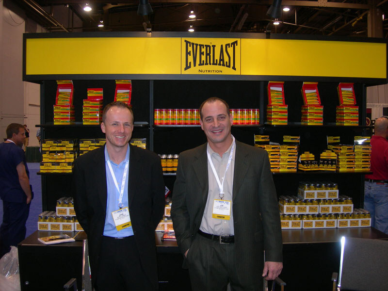 Two men in dark jackets with exhibitor badges stand smiling in front of a yellow Everlast Nutrition sign and black shelves stocked with yellow and red supplement packages and bottles.