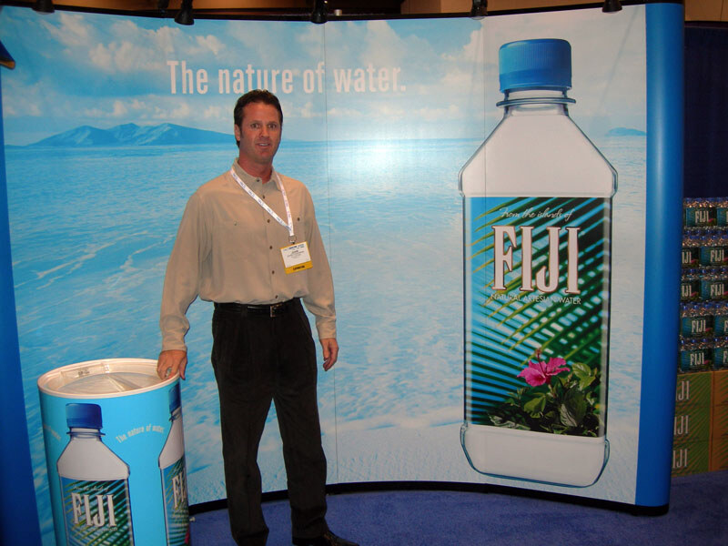 A man in a tan shirt and badge stands before a blue backdrop reading The nature of water, with a large FIJI water bottle graphic and a FIJI cooler beside him.