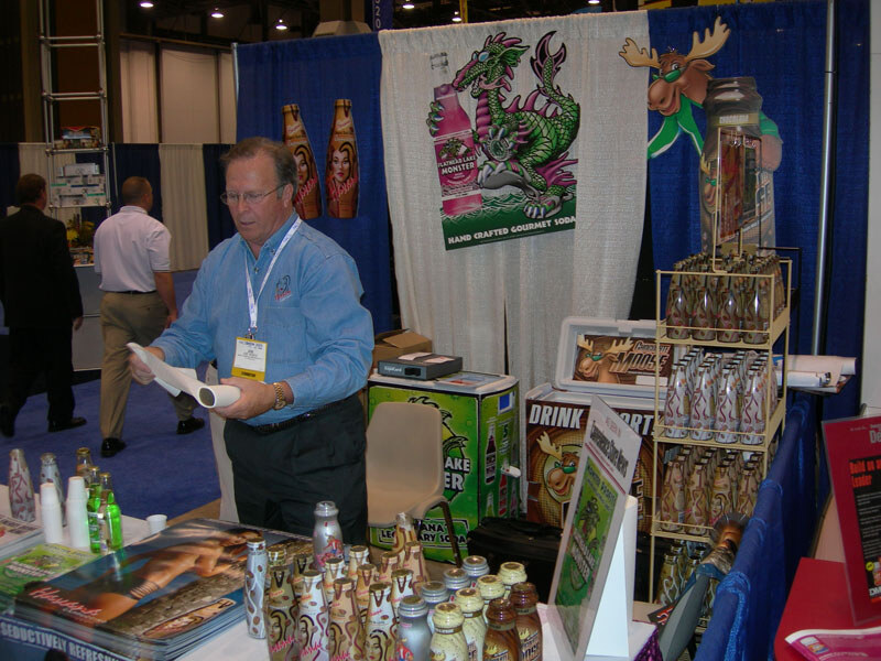 A man in a blue shirt and lanyard looks down at a rolled paper at a booth with Flathead Lake Monster soda banners, a moose graphic, a bottle rack and bottles on the table.