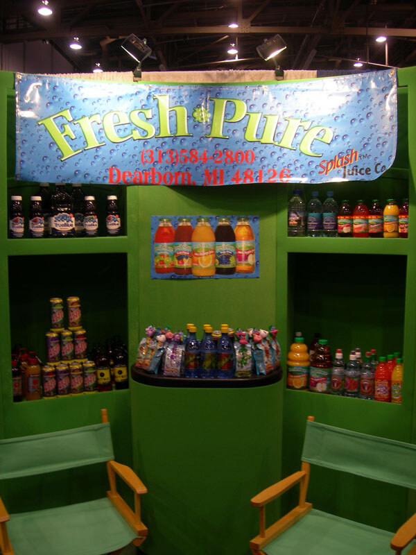 A green booth with a blue Fresh & Pure Splash Juice Co banner, showing shelves of bottled juices and drinks, with two empty green folding chairs in front.