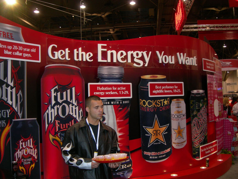A man in a black leather jacket and lanyard holds a tray of small cups in front of a red wall display reading Get the Energy You Want, with large Full Throttle and Rockstar Energy Drink cans.