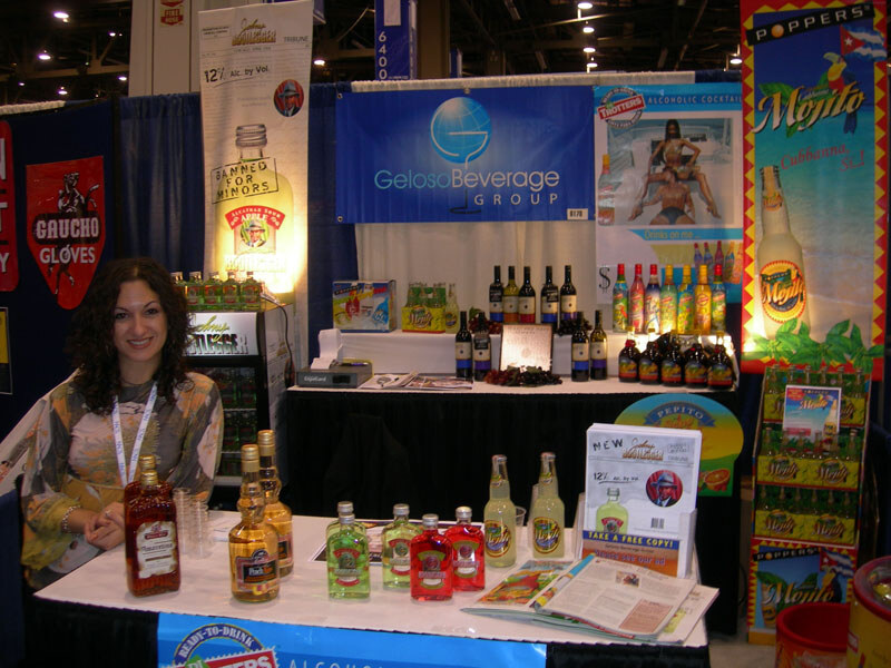 A smiling woman with dark curly hair and a lanyard sits at a table of bottled drinks under a Geloso Beverage Group sign, with Poppers, Trotters and Gaucho Gloves signs around the booth.