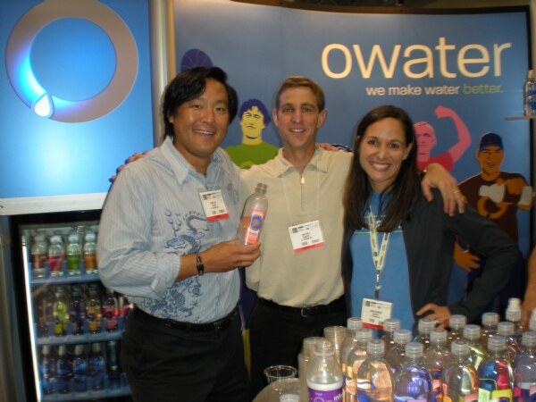 Two men and a woman with arms around each other smile in front of an owater banner. The man on the left holds a water bottle; bottles fill a cooler and the table in front.