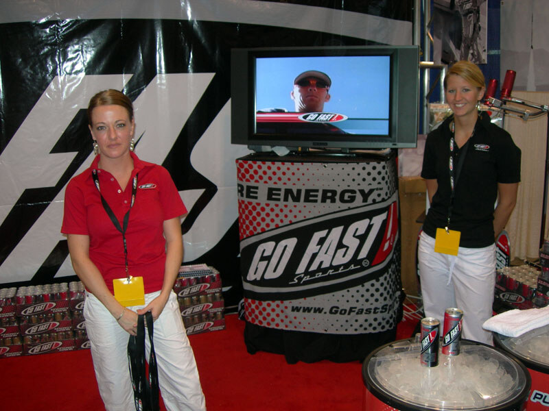 Two women in Go Fast Sports polo shirts and lanyards stand on either side of a Go Fast Sports display with a TV, near cans on ice and stacked cases.