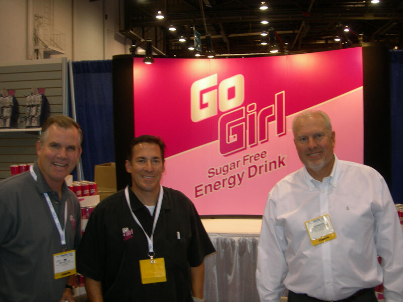Three men with lanyards and badges stand smiling in front of a pink Go Girl Sugar Free Energy Drink backdrop, with stacked red cans and display shelving at left.