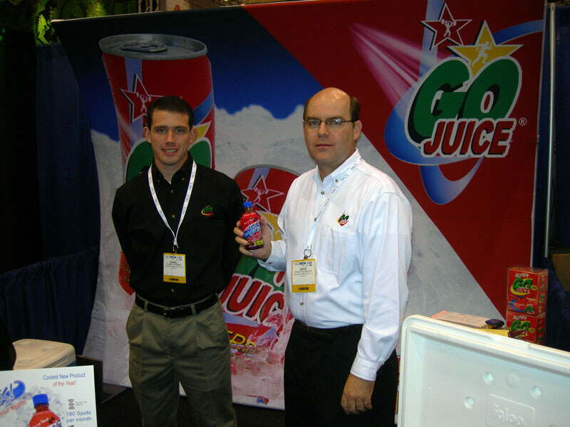 Two men in front of a Go Juice banner; the man on the right, wearing glasses, holds up a bottle of juice. A cooler and Go Juice boxes sit nearby.
