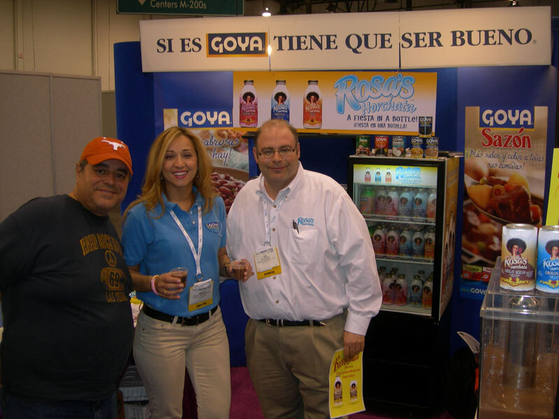 Two men and a woman with blond hair stand in front of a Goya booth with Rosa's Horchata signs, a refrigerated drink cooler and Goya Sazón packaging.