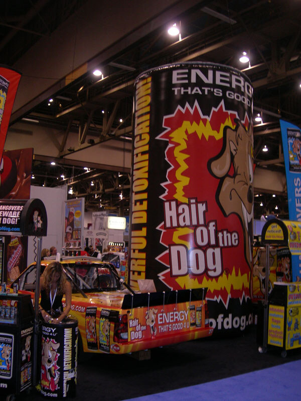 A giant black can-shaped display with a cartoon dog and Hair of the Dog text towers over a decorated orange car; a woman stands at the left near product displays.