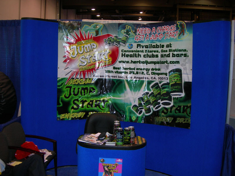 A booth with a blue wall and a green banner reading Herbal Jump Start energy drink; cans sit on a small blue table with a chair and brochures.