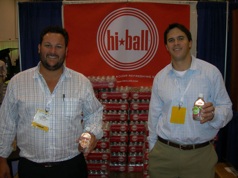 Two men with badges stand in front of a red hi-ball banner and stacked cases of bottled drinks; each holds a bottle.
