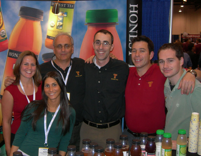 Five men and women in Honest Tea shirts and lanyards pose together in front of large bottle graphics, with bottles of tea on the counter in front.