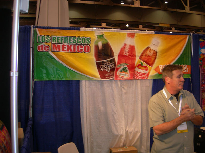 A banner reading "Los Refrescos de México" shows three soda bottles, including Jarritos, above a blue curtain. A man in a green shirt and lanyard stands to the right with hands clasped.