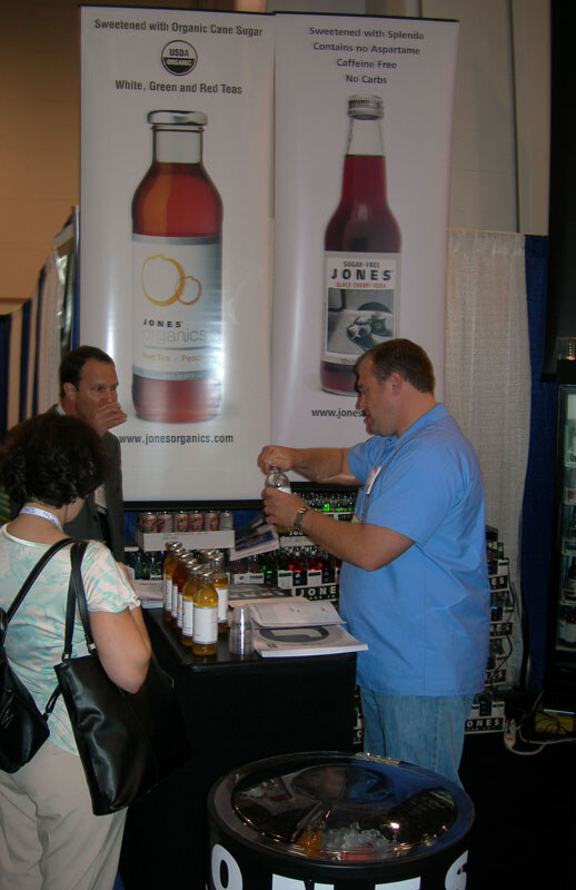 A man in a blue shirt opens a bottle at a booth with Jones Organics and Jones Soda banners; a woman with a bag and another man stand at left.
