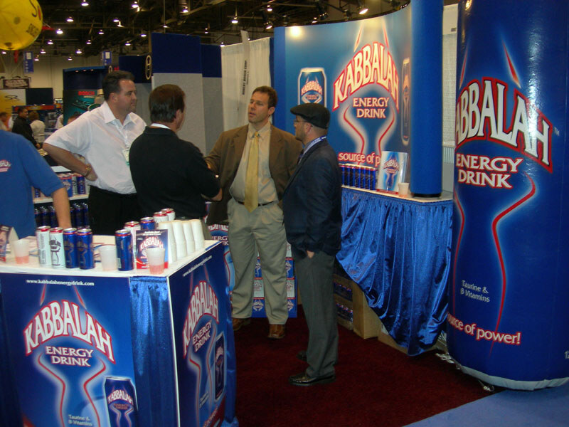 Several men talk at a booth with blue Kabbalah Energy Drink signage, a tall can-shaped column and cans and cups on the counter.