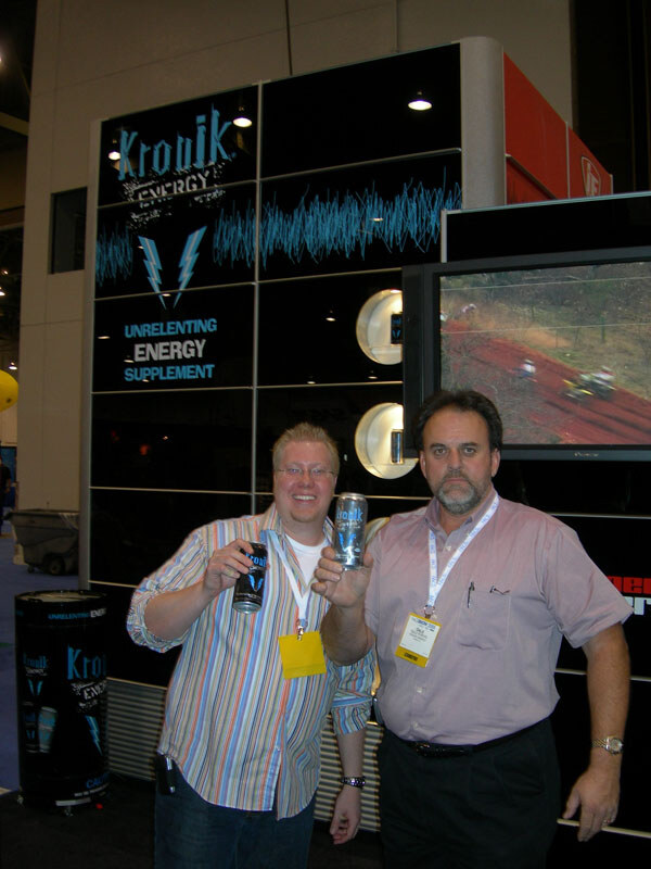 Two men hold up cans in front of a black Kronik Energy display that reads Unrelenting Energy Supplement, with a video screen at right.