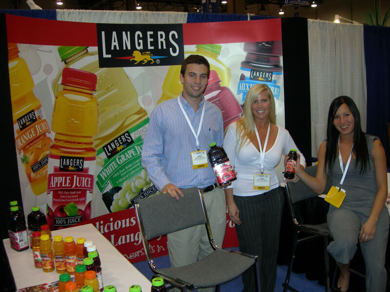 Two men and two women pose in a Langers booth in front of a backdrop of Langers juice bottles, including apple, orange and white grape. Two of them hold bottles of dark red juice.
