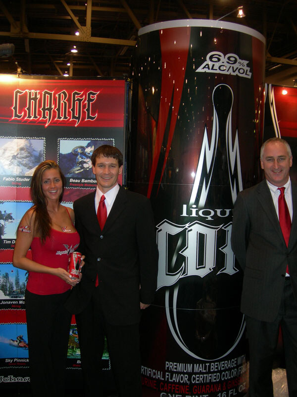 A woman in a red top holding a can and two men in dark suits with red ties stand beside a giant Liquid Charge can display marked 6.9% alc/vol, with a Charge banner behind them.
