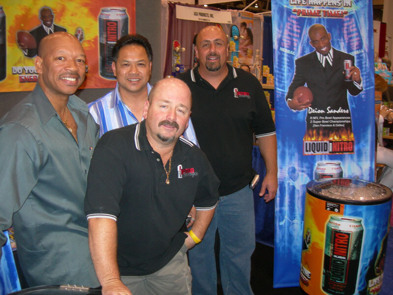 Four men gather close together in a booth with Liquid Nitro banners and a cooler of Liquid Nitro cans. A banner shows a man in a suit holding a football and a can.