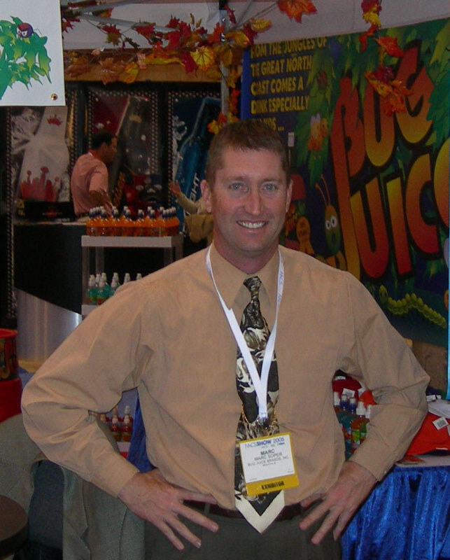 Marc Soper of Bug Juice Brands, Inc. stands smiling with hands on hips, wearing a tan shirt, patterned tie and exhibitor badge, in front of a Bug Juice banner.