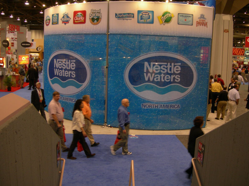A curved blue Nestlé Waters North America booth wall with brand logos including Poland Spring, Ozarka, Zephyrhills and Perrier. Attendees walk past on a blue carpeted aisle.