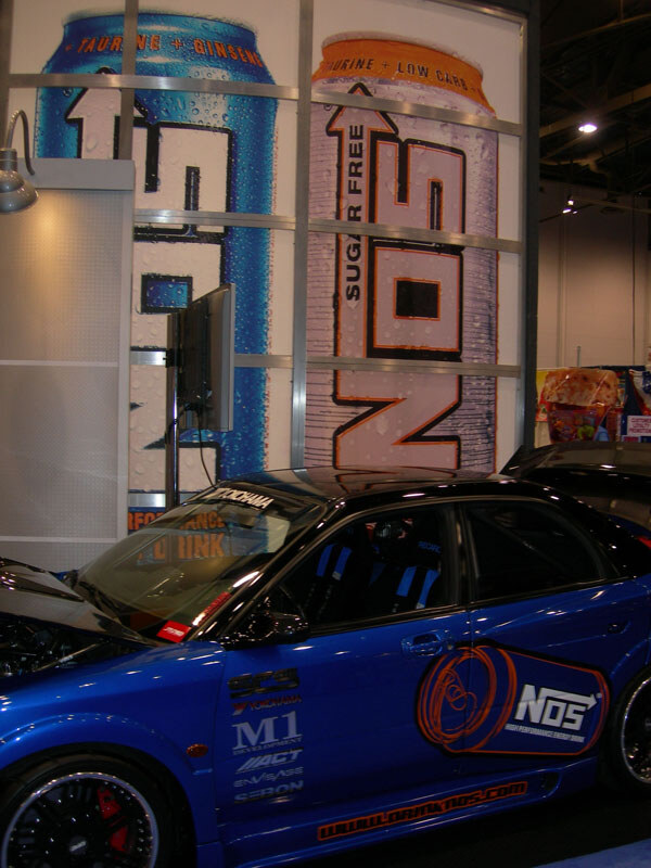 A blue car with NOS and sponsor decals sits in front of a wall graphic of two giant NOS energy drink cans, one regular and one sugar free.