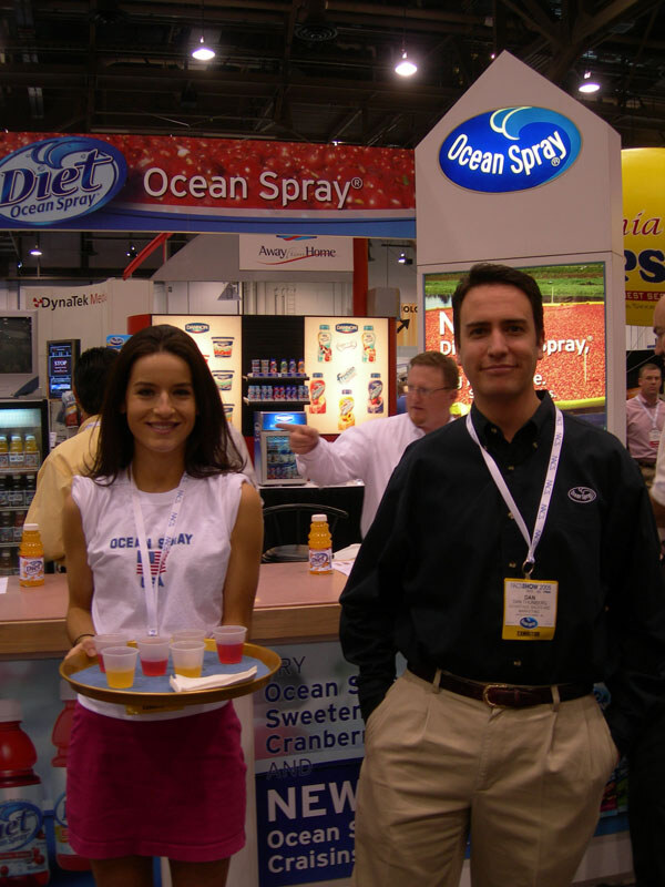 A smiling woman in an Ocean Spray shirt holds a tray of juice samples beside a man in a dark shirt with a badge, in front of Ocean Spray and Diet Ocean Spray signs.