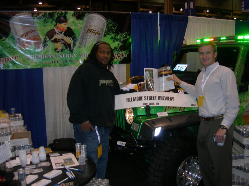 Two men stand beside a green Jeep with a Fillmore Street Brewery sign on its hood, under a Pimp Juice energy drink banner. One man wears a black hoodie, the other a white shirt and holds a magazine.