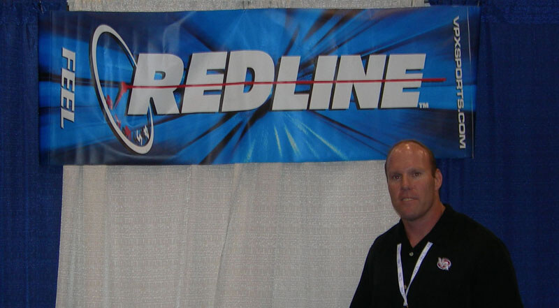 A man in a black polo shirt and lanyard stands in front of a blue Redline banner from VPX Sports, hung on a white curtain between blue booth drapes.