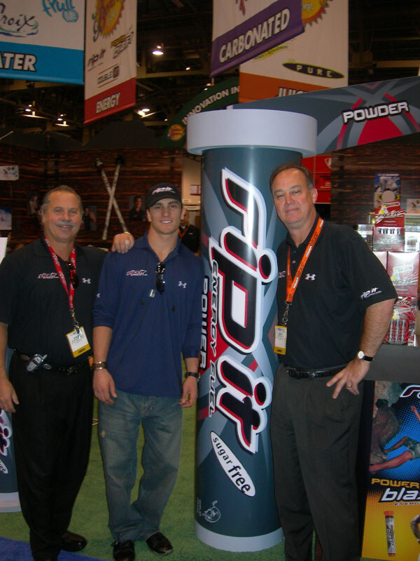 Three men stand around a tall Rip It Energy Fuel display column at a trade show booth. Two wear black Rip It polos with lanyards, and the man between them wears a cap and a blue shirt.