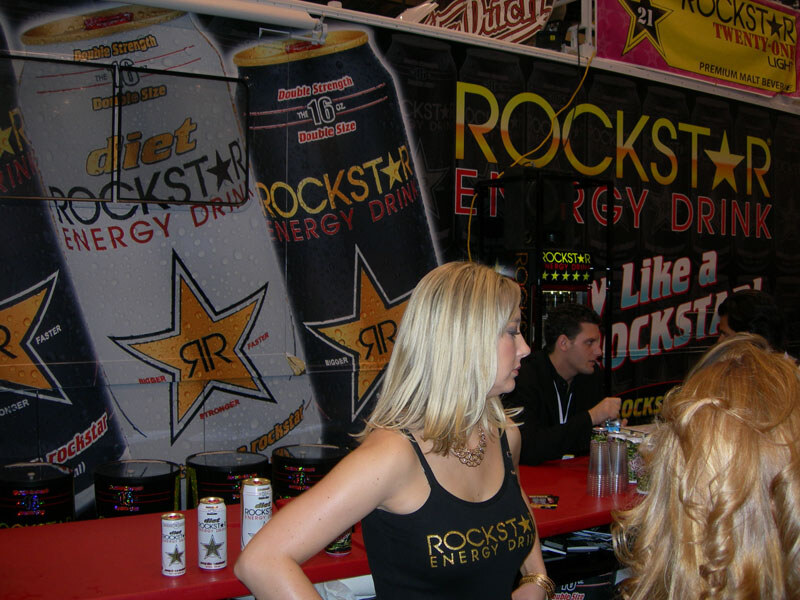 A blonde woman in a black Rockstar Energy Drink tank top stands at a red counter with Rockstar cans in front of large can graphics. A man sits behind the counter in the background.