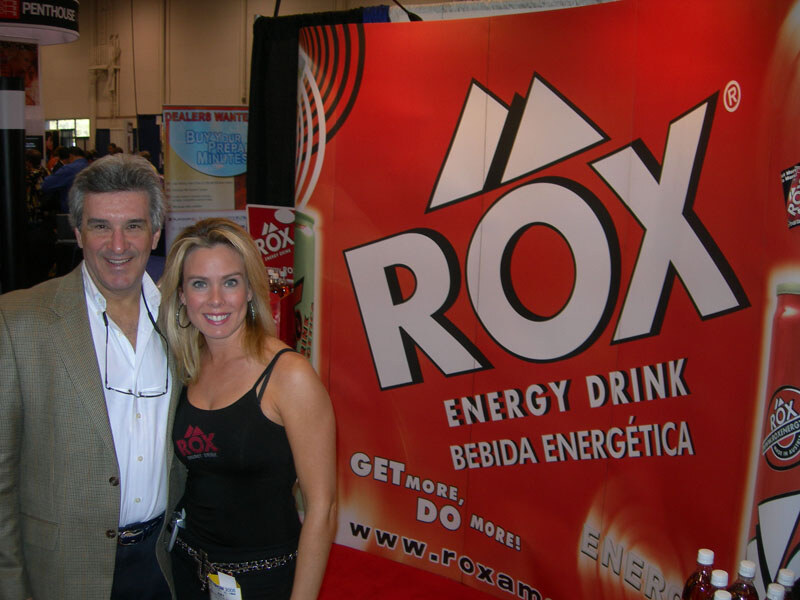 A smiling man in a tan blazer and a smiling woman in a black Rox tank top stand in front of a large red Rox Energy Drink banner reading Get more, do more.