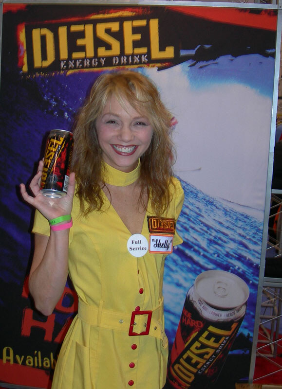 Shelly, wearing a yellow dress with a Diesel patch and name badge, smiles and holds up a can of Diesel Energy Drink in front of a Diesel banner.