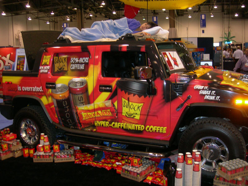 A Hummer wrapped in red and yellow Shock Coffee graphics sits at a trade show booth, with a person sleeping under a blanket on its roof. Shock-A-Lots packages and cups are piled in front.