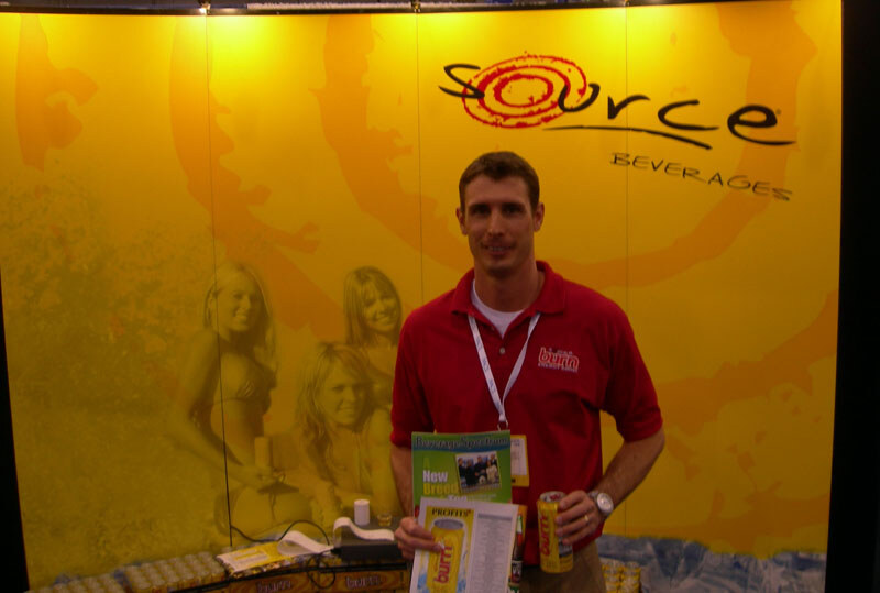 A man in a red Burn polo holds a can of Burn and printed materials in front of a yellow Source Beverages backdrop that shows several women in the background graphic.