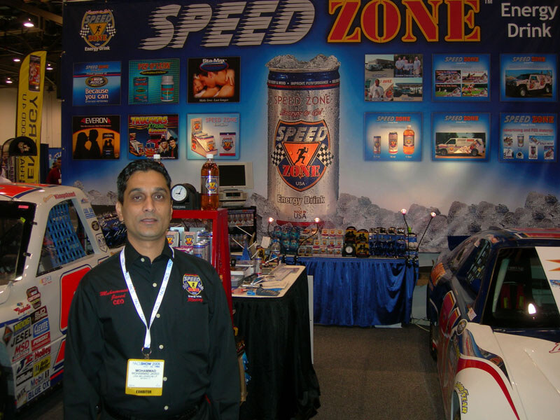 A man in a black Speed Zone shirt and exhibitor badge stands in front of a Speed Zone Energy Drink display with a large can graphic, product tables and race cars on both sides.