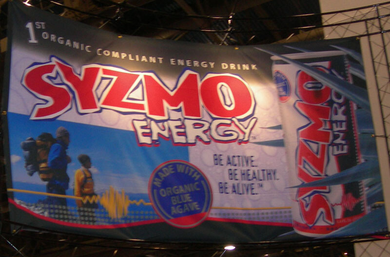 Hanging banner reading Syzmo Energy, 1st Organic Compliant Energy Drink, with a can image, two hikers and a seal reading Made with Organic Blue Agave.