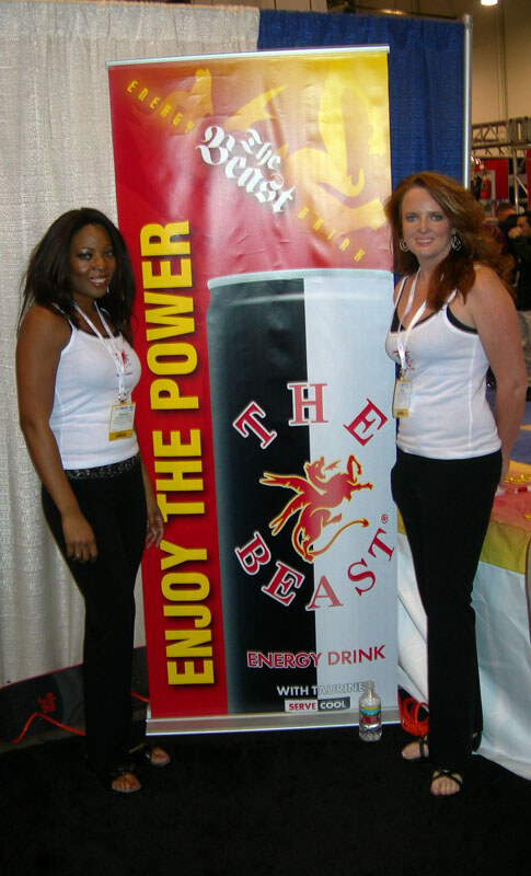 Two women in white tops and black pants with badges stand on either side of a tall The Beast energy drink banner reading Enjoy the Power.