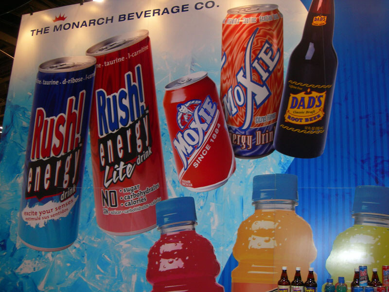 A large Monarch Beverage Co. backdrop shows Rush! Energy cans, Moxie cans, a Dad's Root Beer bottle and colorful juice bottles, with small bottles on a table below.