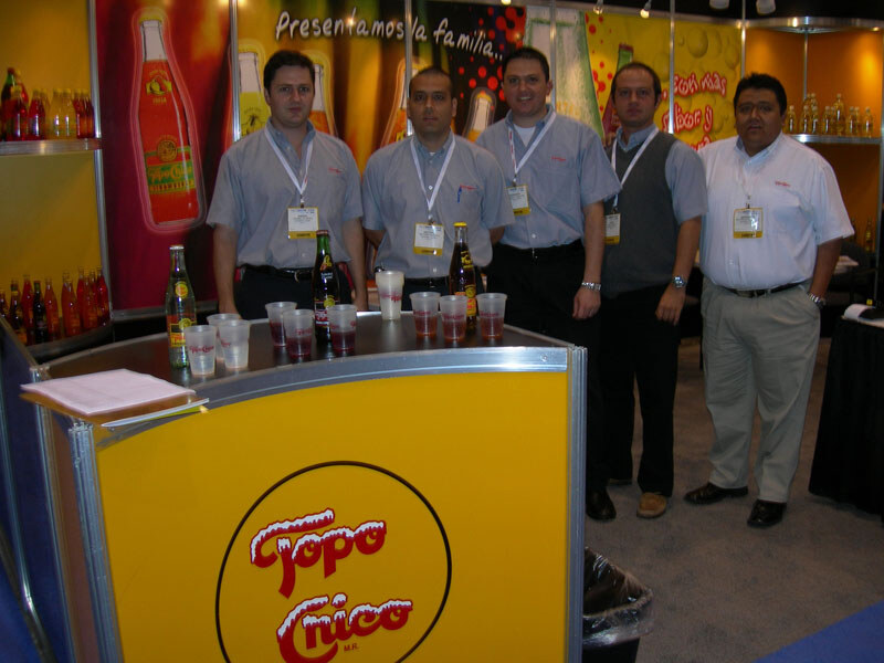 Five men in collared shirts and badges stand behind a yellow Topo Chico booth counter holding bottles and plastic sample cups of a red drink.