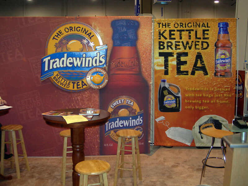 Tradewinds kettle brewed tea booth with large banners showing bottles, a jug and tea bag, a tall wooden table with brochures and several wooden stools.