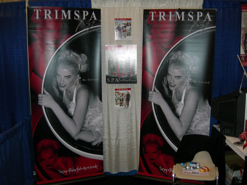 TrimSpa booth with two tall banners showing a blonde woman in black and white, a center panel with product signs, blue curtains and a chair holding items.