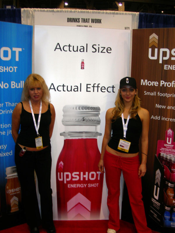 Two women with badges stand before an Upshot energy shot banner reading Actual Size, Actual Effect, under a Drinks That Work sign.