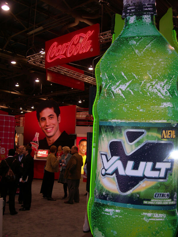 A large green Vault Citrus bottle display stands in the foreground, with a lit Coca-Cola sign and attendees gathered at the Coca-Cola booth behind it.