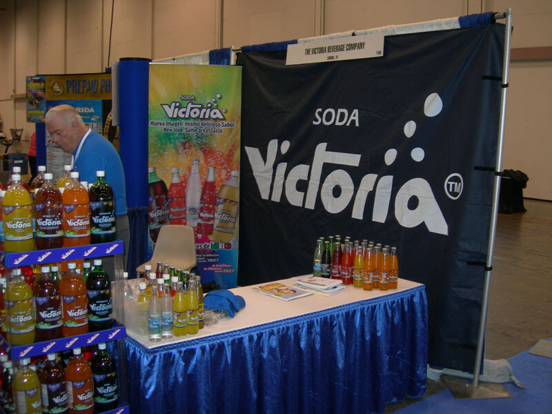 Booth with a dark Soda Victoria banner, a blue-skirted table with bottles and brochures, a shelf of Victoria sodas, and a man in a blue shirt at left.