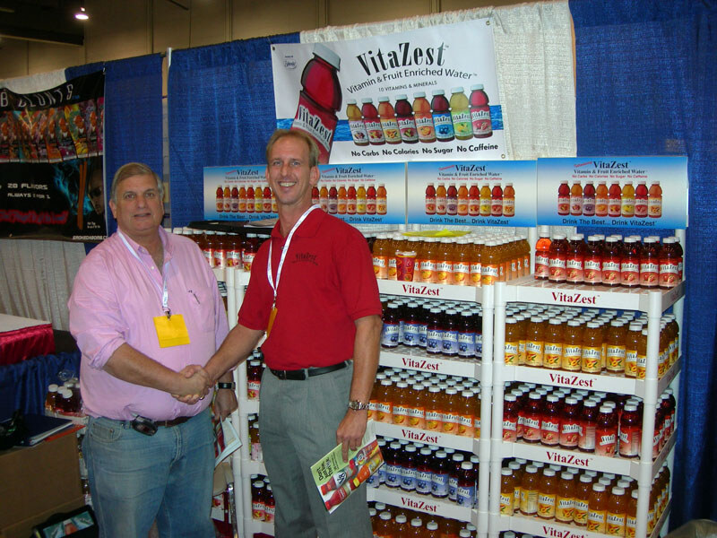Two men shake hands in front of VitaZest vitamin and fruit enriched water shelves and banners; one wears a pink shirt, the other a red VitaZest polo.
