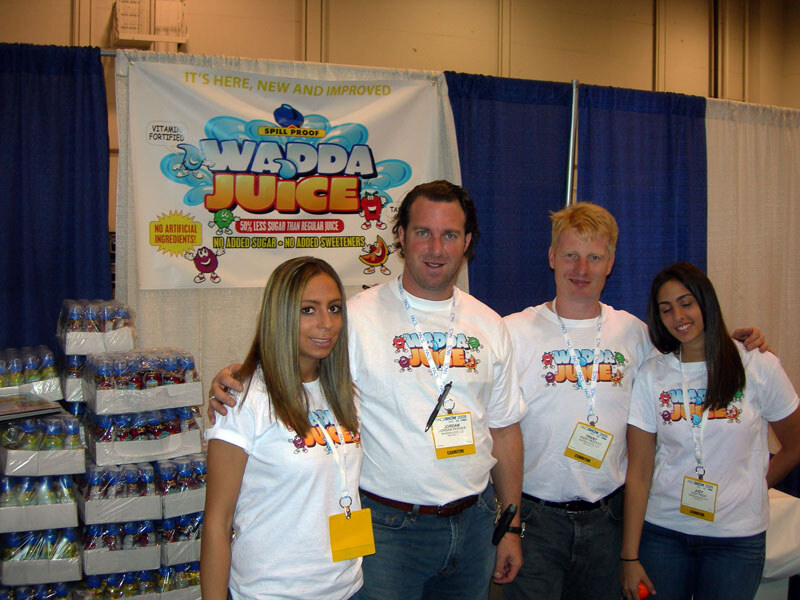 Four people in white Wadda Juice T-shirts and badges pose in front of a Wadda Juice banner and stacked cases of bottled juice.