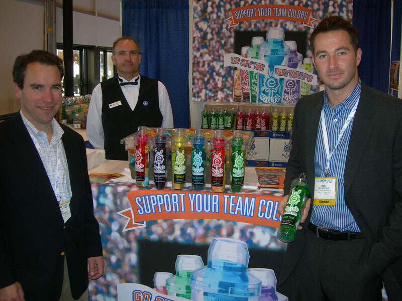 Three men stand at a Wet Willy's booth with rows of colorful bottles and a Support Your Team Colors banner; the man at right holds a green bottle and wears a badge.