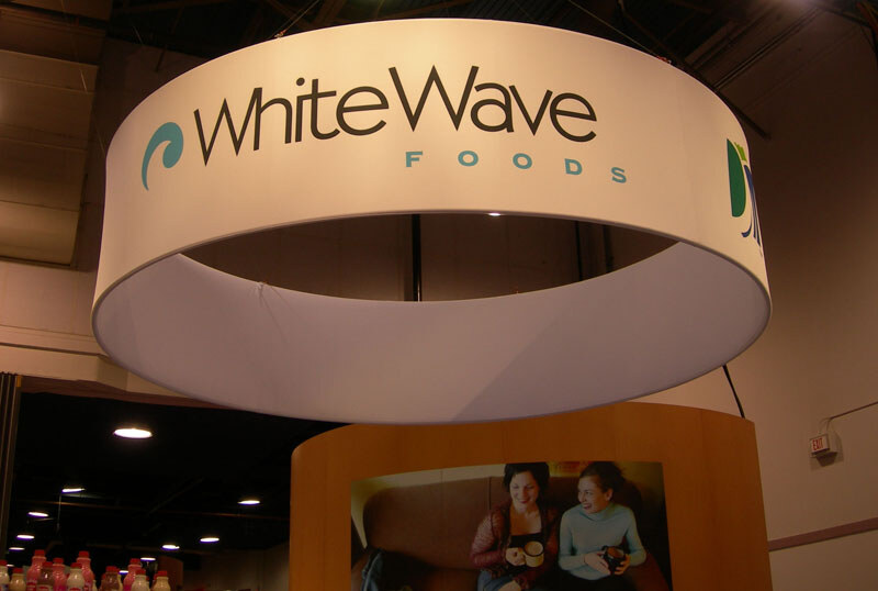 A large white circular hanging sign reading WhiteWave Foods hangs from the ceiling above a curved wooden booth wall with a photo of two smiling women holding mugs.