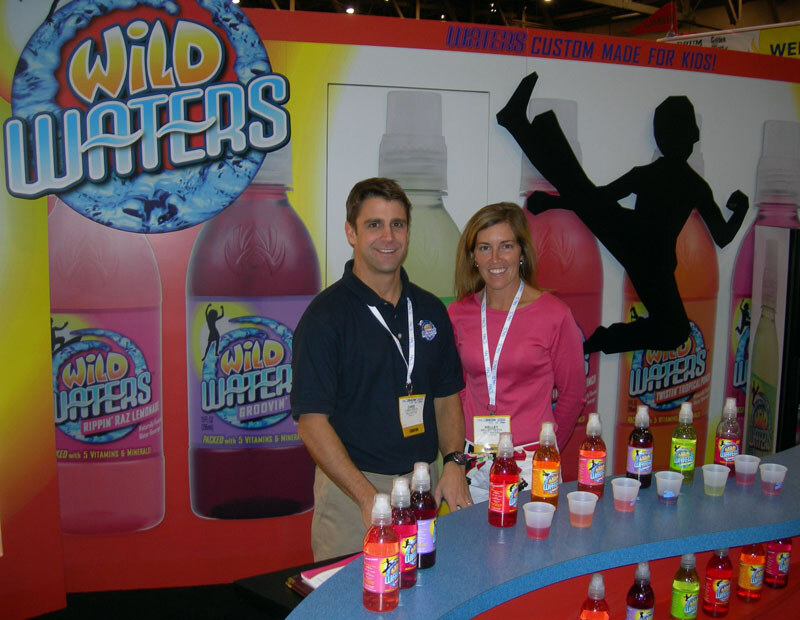 A man in a navy polo and a woman in a pink top, both wearing lanyards, stand smiling behind a counter of Wild Waters bottles and sample cups in front of a Wild Waters backdrop.